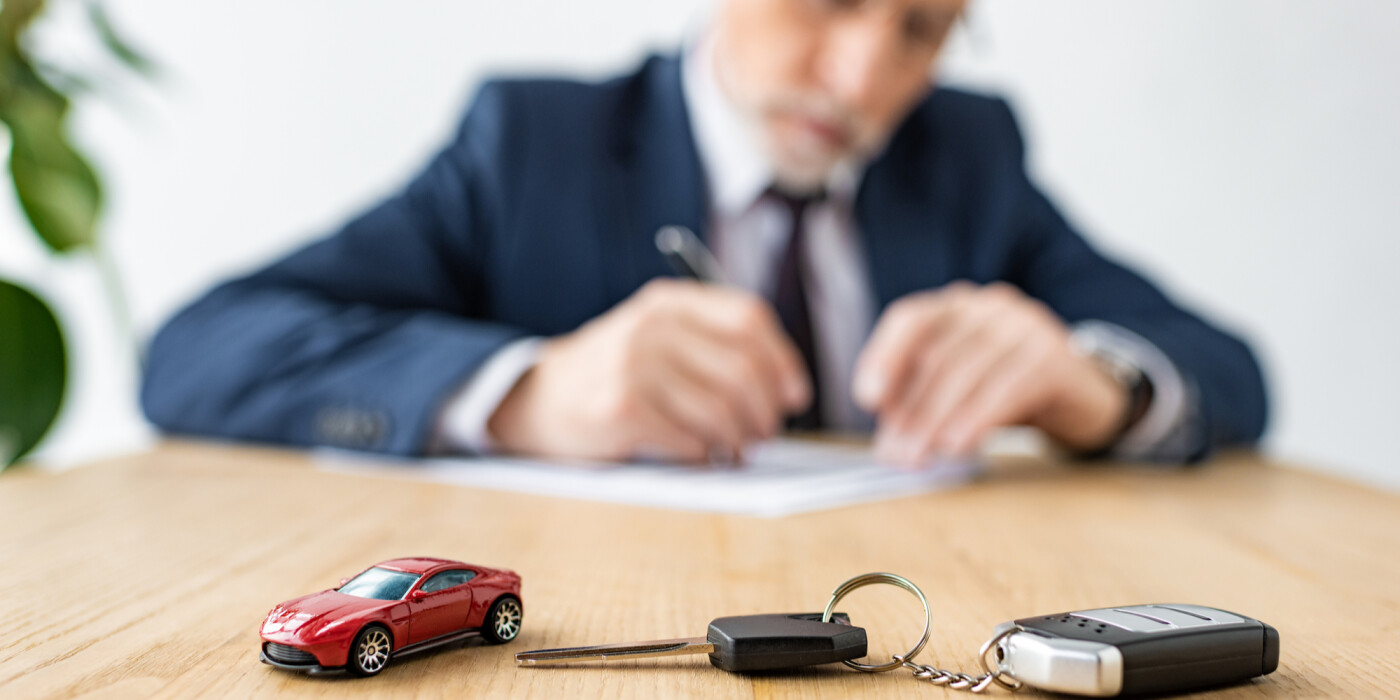 Car keys and small toy car on a desk with man signing paperwork in the background