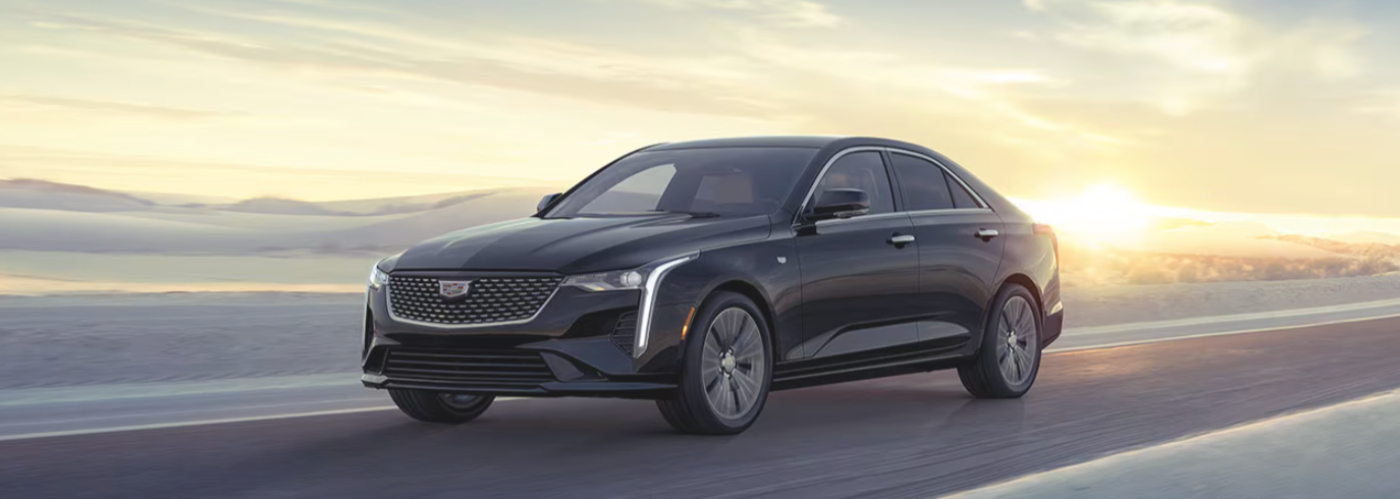 The black CT4 is driving along the ocean with the sunset behind it. 